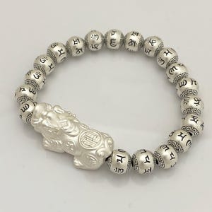 May include: Silver bracelet featuring a detailed Pixiu charm and round beads with engraved symbols. The bracelet is strung on an elastic band. The Pixiu charm is a mythical creature, often associated with attracting wealth and good fortune.