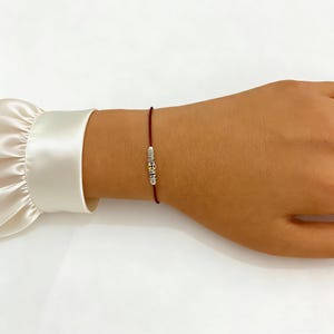 May include: A red string bracelet with a silver charm. The charm has a detailed design. The bracelet is worn on a wrist with a white satin sleeve in the background. The bracelet is a simple, elegant piece of jewelry.