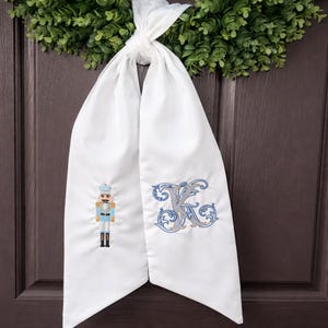 Puede incluir: Bufanda de puerta de tela blanca con un soldado cascanueces bordado en azul y dorado y una letra monograma "K" azul y gris. La bufanda está atada en la parte superior y cuelga contra una puerta marrón oscuro.
