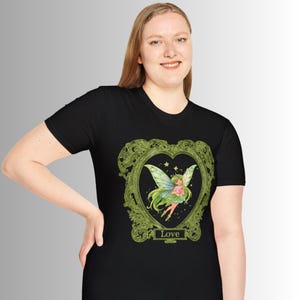 May include: Black t-shirt featuring a green heart-shaped frame with a fairy illustration. The fairy has green hair and wings, holding a pink flower. The word "Love" is printed at the bottom of the frame.