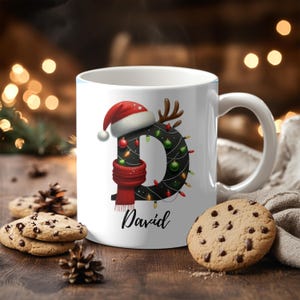 Pode incluir: Caneca de cerâmica branca com a letra "D" preta decorada com luzes de Natal, um gorro de Papai Noel e um cachecol vermelho. O nome "David" está escrito abaixo. Biscoitos com gotas de chocolate e pinhas estão na superfície de madeira.