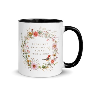 May include: White ceramic mug with a black interior and handle. The mug features a floral wreath design with pink and yellow flowers, a small bird, and the text "THOSE WHO WISH TO SING ALWAYS FIND A SONG."