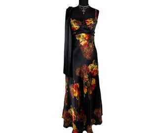 Puede incluir: Un vestido de noche de satén negro con un estampado floral en tonos rojos, amarillos y naranjas. El vestido presenta un escote corazón, tirantes finos y una falda larga y fluida. Un chal negro está drapeado sobre el hombro.