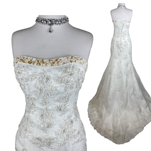 May include: A strapless white wedding dress featuring intricate floral lace embroidery, clear beadwork, and a gold sequined trim along the neckline. The gown has a fitted bodice and a flared skirt with a train, complemented by a crystal choker necklace.