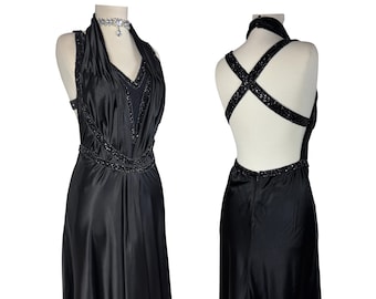 May include: A black halter dress with a beaded neckline and waist, displayed on a mannequin. The dress features a low back with beaded straps. The fabric has a satin sheen. A silver necklace is around the mannequin's neck.