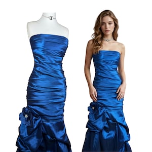 Puede incluir: Un vestido palabra de honor en azul real con cuerpo fruncido y silueta entallada. La falda incluye un detalle fruncido lateral con una gran flor de tela en la cadera, que se transforma en un bajo acampanado con acabado satinado.