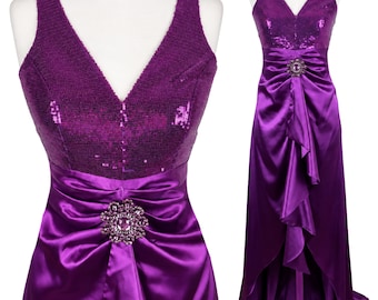 May include: A floor-length purple dress featuring a sleeveless bodice covered in matching sequins with a V-neckline. The waist is accented by a circular jeweled brooch, and the skirt has a satin finish with a cascading ruffled front detail.