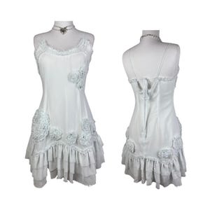 Puede incluir: Vestido blanco con tirantes finos, adornos florales y bajo con volantes. El vestido presenta tirantes finos y un delicado ribete de encaje en el escote. Se muestra en un maniquí, mostrando la vista frontal y trasera.