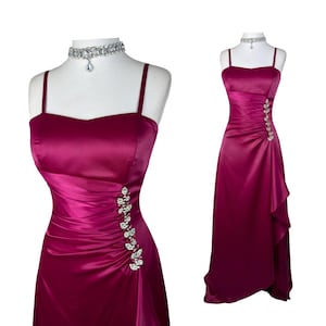 May include: A floor-length magenta satin gown featuring thin shoulder straps, a sweetheart neckline, and side-ruched detailing accented with a vertical row of clear rhinestones. The dress is displayed on a mannequin wearing a matching rhinestone choker.