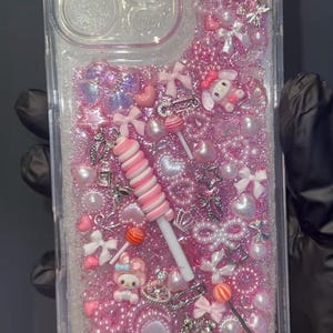 May include: Clear phone case filled with pink glitter, pearls, bows, and charms. Features a large pink flower, a lollipop, and a My Melody character. The case is designed with a clear, protective bumper.