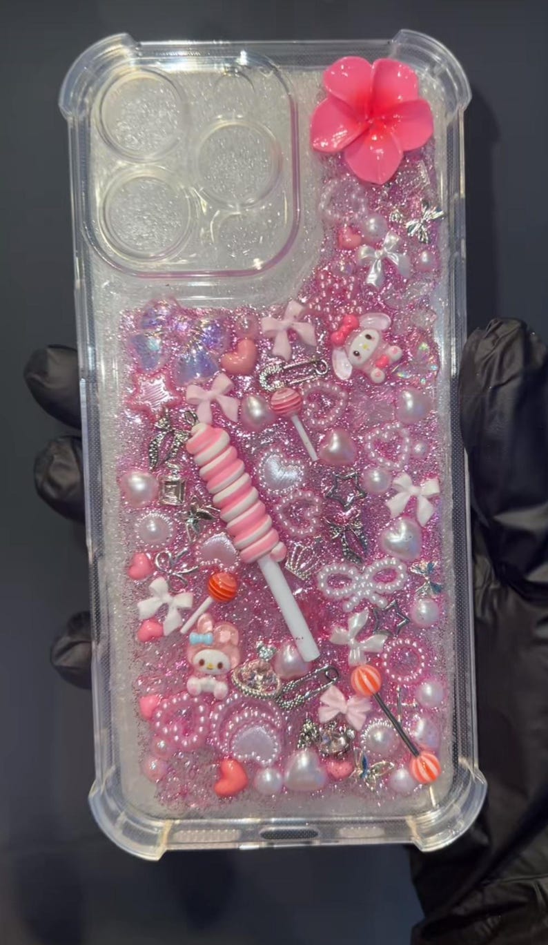 May include: Clear phone case filled with pink glitter, pearls, bows, and charms. Features a large pink flower, a lollipop, and a My Melody character. The case is designed with a clear, protective bumper.