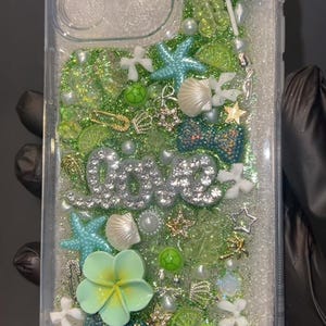May include: Clear phone case with a glittery, ocean-themed design. Features a bow, starfish, seashells, and the word "love" in silver rhinestones. Includes green and blue accents, and a light green flower.