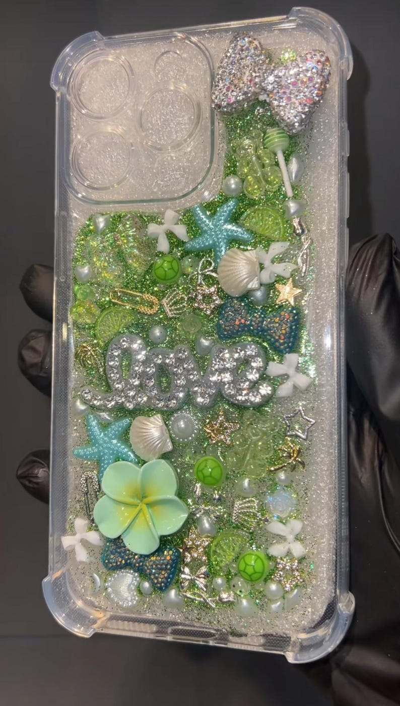 May include: Clear phone case with a glittery, ocean-themed design. Features a bow, starfish, seashells, and the word "love" in silver rhinestones. Includes green and blue accents, and a light green flower.