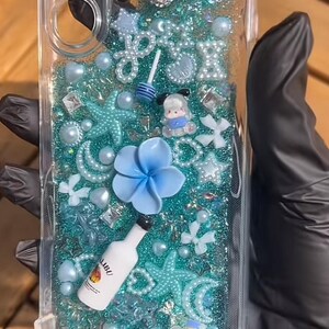 May include: Clear phone case with a turquoise glitter background, adorned with various embellishments. Features a large, jeweled bow, blue flower, starfish, pearls, and a miniature Malibu bottle. The case is designed for a phone.