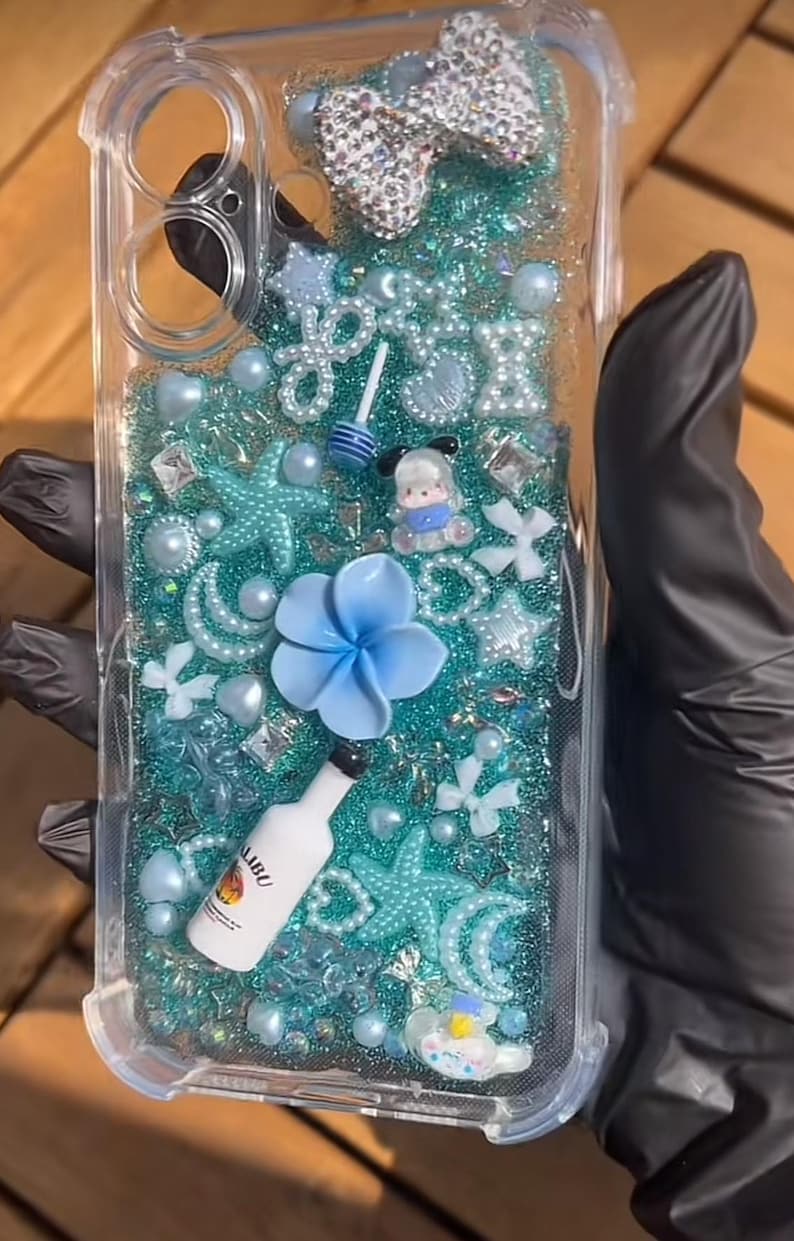 May include: Clear phone case with a turquoise glitter background, adorned with various embellishments. Features a large, jeweled bow, blue flower, starfish, pearls, and a miniature Malibu bottle. The case is designed for a phone.