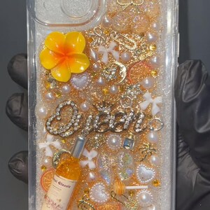 May include: Clear phone case with a glittery, decorative interior. Embellishments include a yellow flower, pearls, gold charms, and the word "Queen" in rhinestones. A small bottle with text is also included.