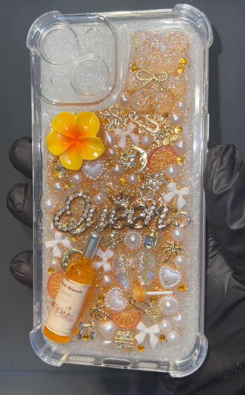 May include: Clear phone case with a glittery, decorative interior. Embellishments include a yellow flower, pearls, gold charms, and the word "Queen" in rhinestones. A small bottle with text is also included.