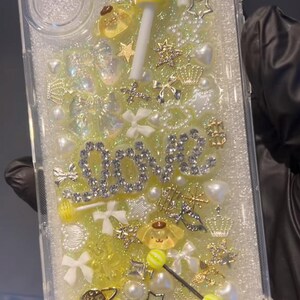 May include: Clear phone case with a glittery yellow and white design. Embellishments include a yellow lollipop, the word "LOVE" in silver, stars, bows, pearls, and gold charms. The case is filled with small decorative elements.