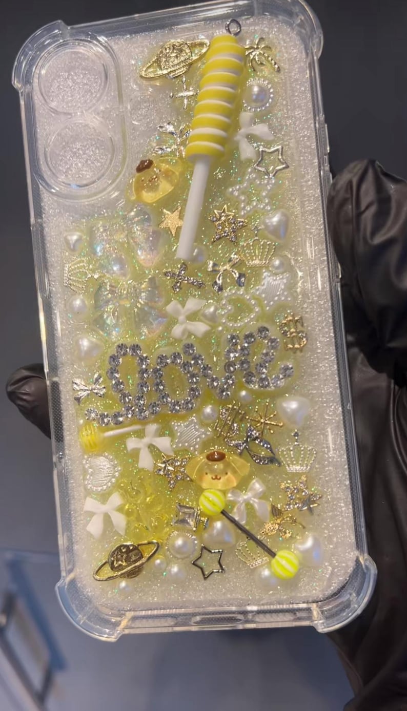May include: Clear phone case with a glittery yellow and white design. Embellishments include a yellow lollipop, the word "LOVE" in silver, stars, bows, pearls, and gold charms. The case is filled with small decorative elements.