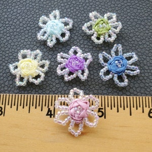 May include: Six handmade floral embellishments. Each flower features a fabric center in various colors, including blue, green, yellow, purple, and pink. The petals are outlined with clear beads, creating a delicate, decorative effect. The flowers are arranged on a dark surface.