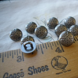 May include: A collection of antique metal buttons with a fleur-de-lis design. The buttons are a dark, tarnished silver color and are arranged on a white floral patterned fabric next to a wooden ruler. The ruler is marked in inches.