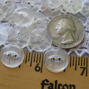 May include: A collection of clear, round buttons with two holes, alongside a US quarter coin for size comparison. The buttons are translucent and appear to be made of plastic. A wooden ruler with the word "falcon" is also visible.