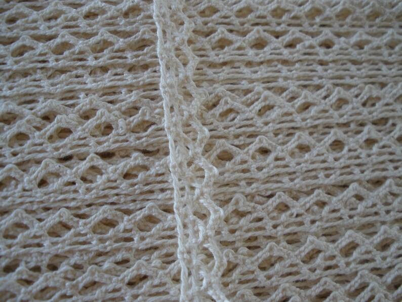 May include: Close-up of a white crocheted fabric with a diamond pattern. The fabric has a delicate, lacy texture.