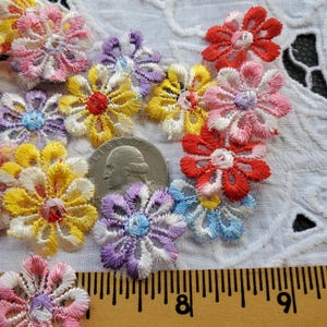 May include: Assortment of colorful embroidered flower appliques in various shades of pink, red, yellow, blue, and purple. The appliques are approximately 1 inch in diameter and are arranged on a white fabric background.