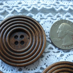 May include: Close-up of large, dark brown wooden buttons with concentric circle design. Each button has four small holes. A US quarter coin is next to the button for size comparison. The quarter is dated 1980.