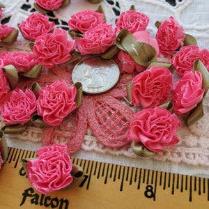 May include: A collection of small, pink fabric roses with green ribbon accents. A U.S. quarter coin is included for size reference. The roses are scattered on a lace-like fabric surface, with a ruler in the foreground.
