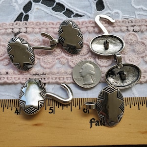 May include: Five silver-toned metal buttons with a star design and a hook closure. The buttons are oval shaped and have a textured surface. The buttons are arranged on a white lace surface with a ruler in the background.