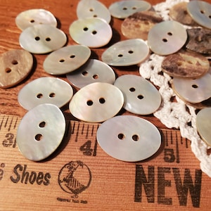 May include: A collection of oval-shaped, iridescent mother-of-pearl buttons with two holes each. The buttons are arranged on a wooden surface with a ruler and a piece of lace.