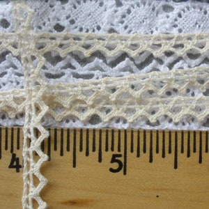 May include: Close-up of a white and cream lace trim with a scalloped edge. The lace is measured against a wooden ruler with markings in inches.