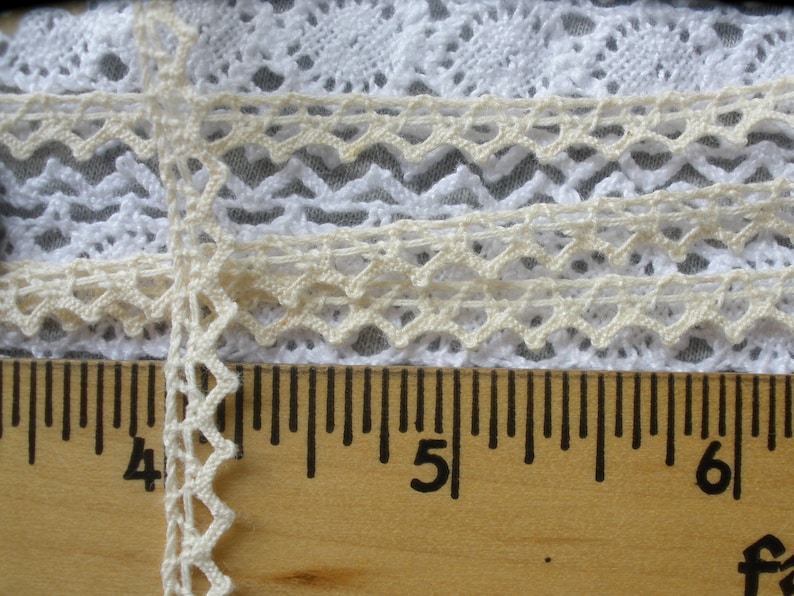 May include: Close-up of a white and cream lace trim with a scalloped edge. The lace is measured against a wooden ruler with markings in inches.