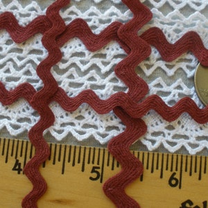 May include: Close-up of a length of burgundy ric rac trim, with a zig-zag pattern. The trim is placed on a white lace background. A ruler and a quarter are also in the image, providing scale.