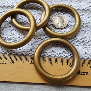 May include: Four brass rings, each with a diameter of approximately 2 inches, are arranged on a white lace fabric. A ruler is placed below the rings, with the numbers 2, 3, 4, 5, 6, and 7 visible.