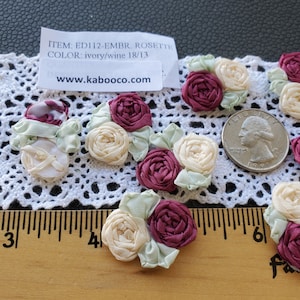 May include: A close-up of ivory and burgundy fabric rosettes. The rosettes are about 1 inch in diameter and are arranged in a cluster on a white lace surface. The text "ITEM: ED112-EMBR. ROSETTE COLOR: ivory/wine 18/13 www.kabooco.com" is visible in the image.