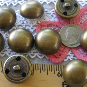 May include: A collection of antique brass-colored dome-shaped buttons. Some buttons have a shank and holes for sewing. A ruler and a quarter are included for size reference. The buttons are arranged on a lace and floral background.
