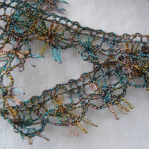 May include: Close-up of a metallic lace trim with a teal, gold, and brown color scheme. The lace has a delicate, intricate pattern and a frayed edge.