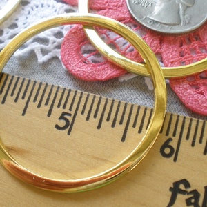 May include: Close-up of several gold-colored metal rings, possibly for jewelry making or crafts. A wooden ruler with inch markings is in the foreground, with the rings overlapping a pink crocheted doily and a silver coin. The rings are approximately 2 inches in diameter.