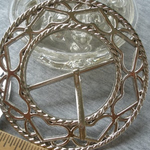 May include: Silver metal belt buckle with a decorative, intricate design. The buckle is circular in shape with a rope-like border and a delicate, openwork pattern.