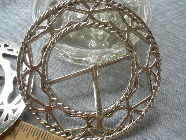 May include: Silver metal belt buckle with a decorative, intricate design. The buckle is circular in shape with a rope-like border and a delicate, openwork pattern.