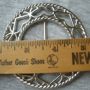 May include: Silver metal belt buckle with a decorative design of interlocking circles and hearts. The buckle is approximately 5 cm in diameter.