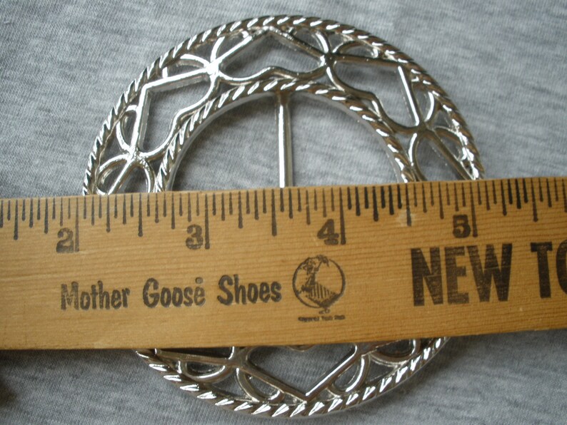 May include: Silver metal belt buckle with a decorative design of interlocking circles and hearts. The buckle is approximately 5 cm in diameter.