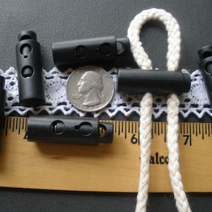 May include: Six black plastic cord locks with a white cord running through them. The cord locks are about 1 inch long and have a small hole in the center. The cord locks are sitting on a white lace fabric and a wooden ruler.