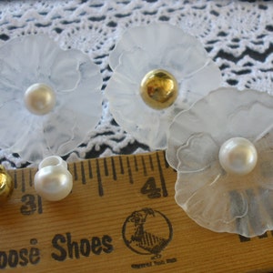 May include: Four white plastic flower-shaped buttons with a pearl-like center. The buttons are arranged on a white lace fabric with a wooden ruler in the foreground.