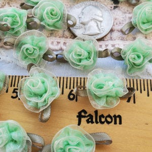 May include: A close-up of a collection of green and white fabric flower embellishments. The flowers are arranged in a row on a wooden surface with a ruler and the word "Falcon" printed on it.