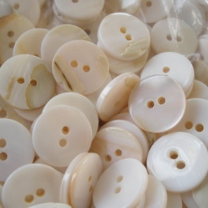 May include: A close-up of a pile of white and cream-colored round buttons with two holes each. The buttons are made of a smooth, shiny material and have a slightly iridescent sheen.