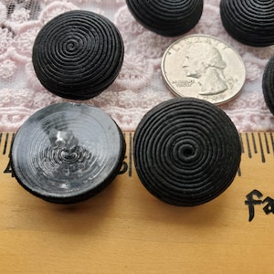 May include: A collection of black buttons with a spiral design. The buttons are round and have a textured surface. A U.S. quarter is included for size reference. The buttons are on a wooden surface.