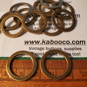 May include: A pile of 10 brass jump rings, approximately 1 inch in diameter, on a white surface with a business card that reads "www.kabooco.com Vintage buttons, supplies & cool finds too!"